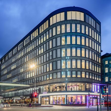 Yotel Glasgow Luaran gambar The photo shows a modern office building with a distinctive curved shape and a facade made of large glass windows. It is illuminated against a twilight sky, giving it a contemporary appearance. The ground floor features large glass entrance areas, li