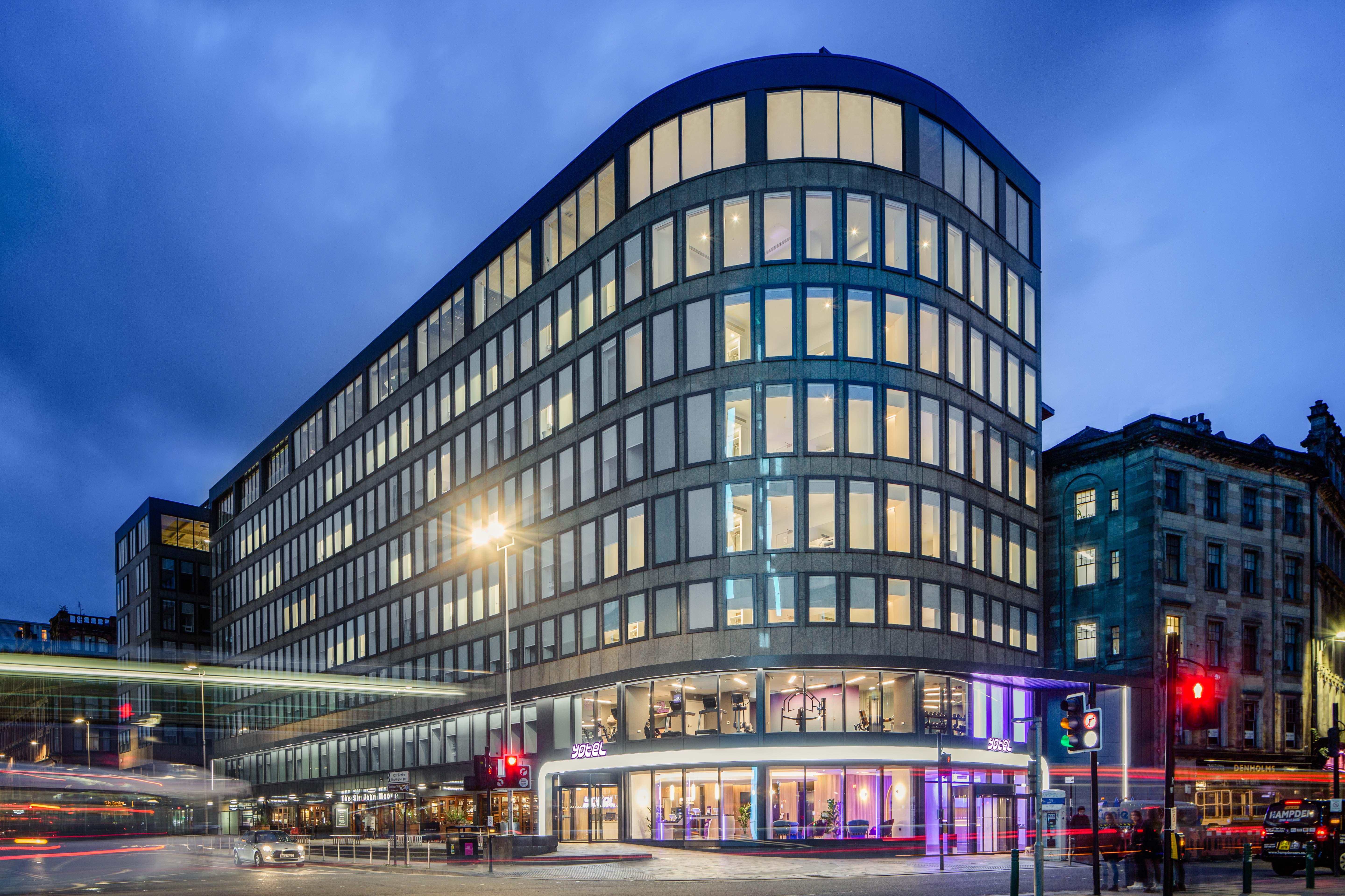 Yotel Glasgow Luaran gambar The photo shows a modern office building with a distinctive curved shape and a facade made of large glass windows. It is illuminated against a twilight sky, giving it a contemporary appearance. The ground floor features large glass entrance areas, li